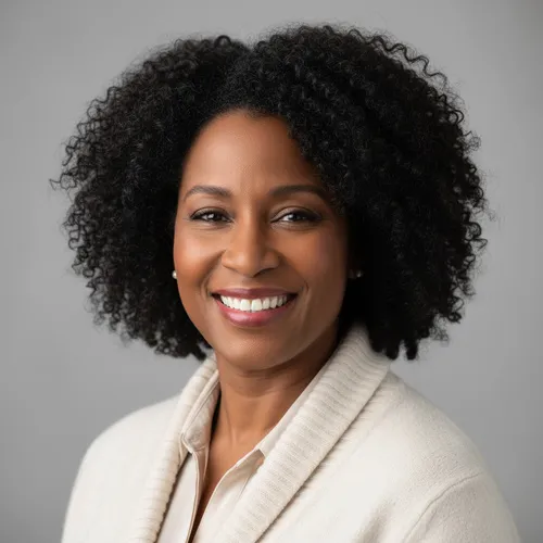 A woman with curly hair and a cream sweater smiles at the camera against a plain gray background.
