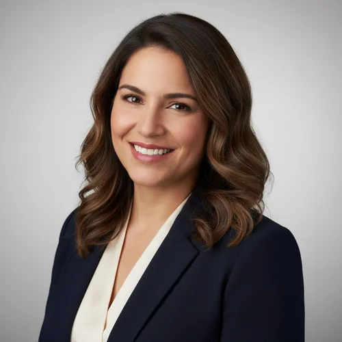 A woman with shoulder-length brown hair, wearing a dark blazer over a light blouse, smiles in front of a plain gray background.