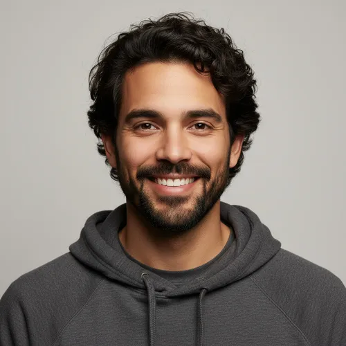 A man with dark curly hair and a beard is smiling at the camera, wearing a gray hoodie against a plain light background.