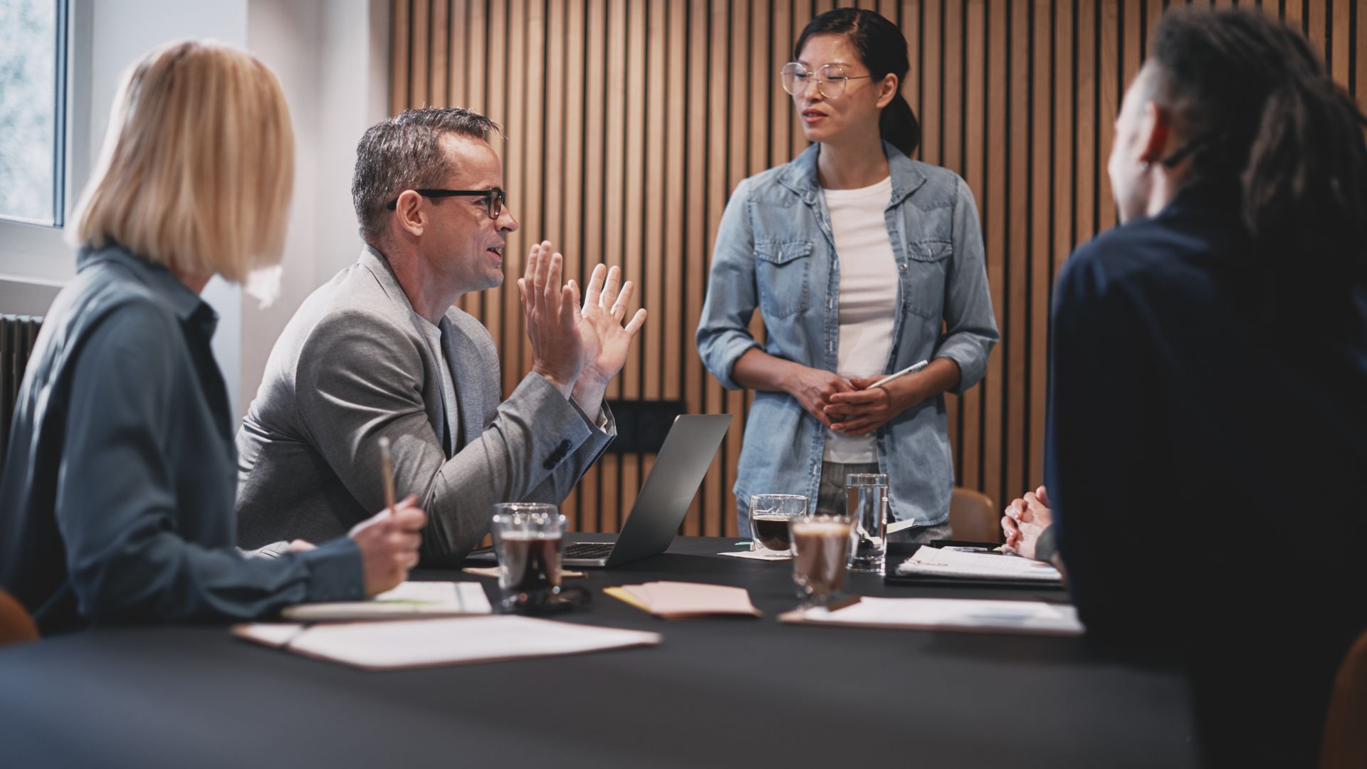 Four people sit around a conference table with documents and coffee, while one woman stands and speaks to the group in a modern meeting room.