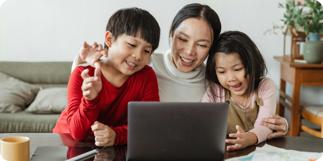 An adult and two young children sit together at a table, smiling and looking at a laptop screen in a home setting.