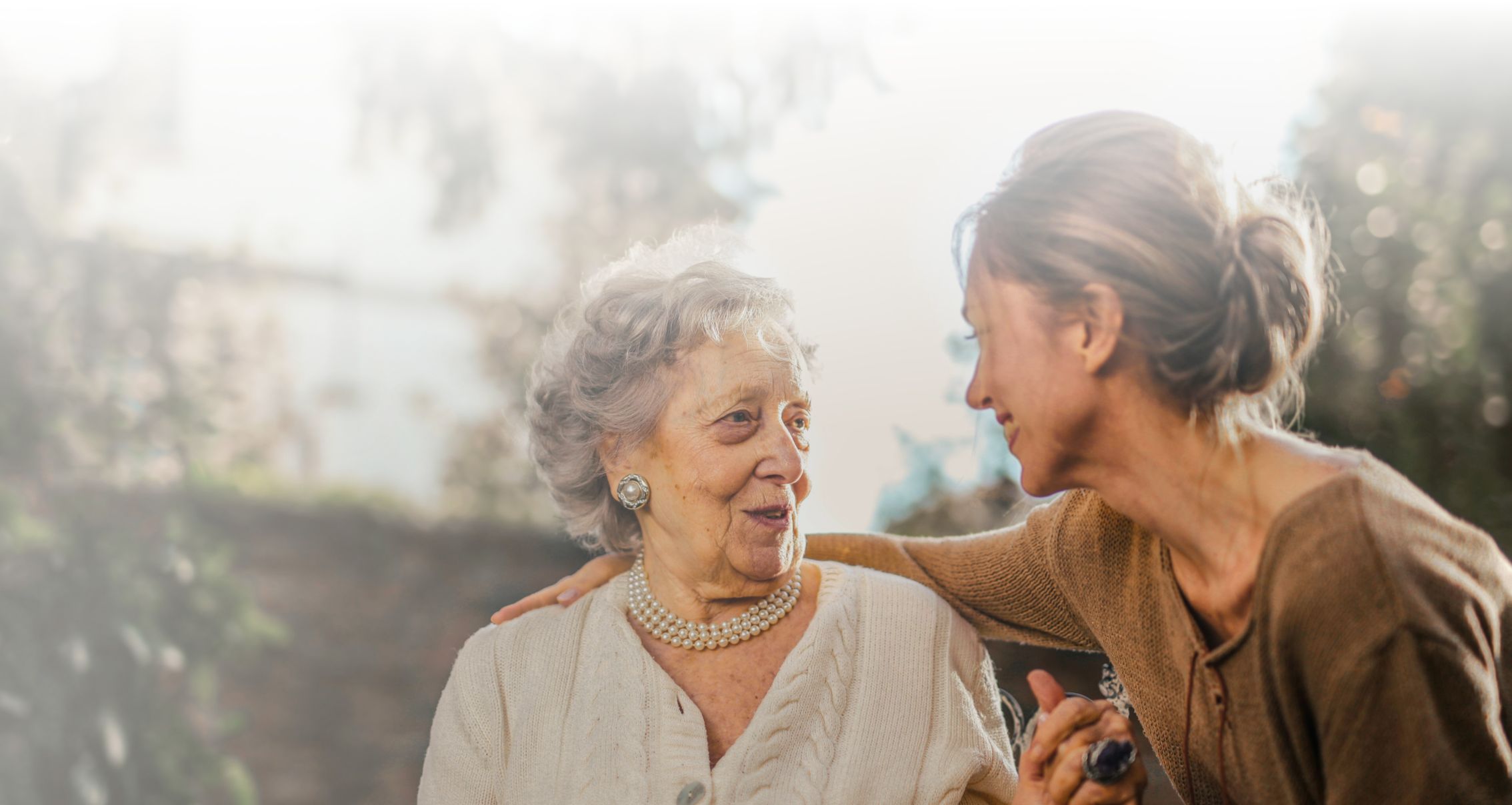 An older woman and a younger woman sit outside, smiling at each other, with the younger woman resting her arm on the older woman's shoulder.