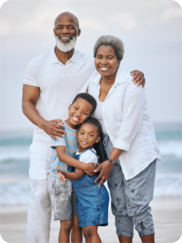 A smiling family of four poses together on a beach, with two adults standing behind two children who are hugging in front of them. The ocean is visible in the background.