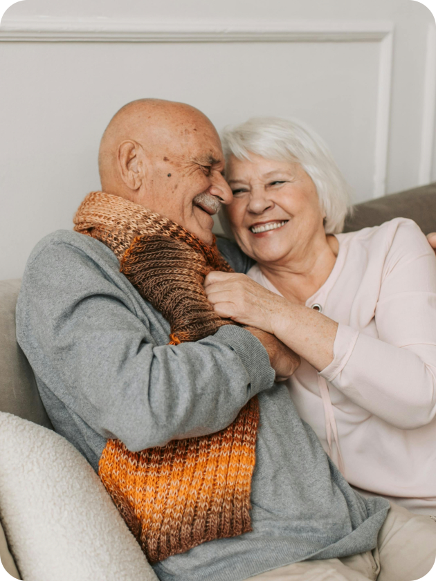 An elderly man and woman sit close together on a couch, smiling and embracing. The man wears a grey sweater and orange scarf; the woman wears a light pink top.