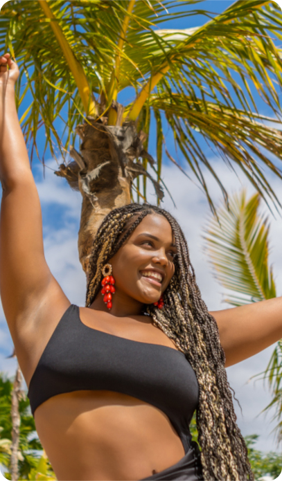 Woman with long braids in a black swimsuit smiles with arms raised beneath palm trees on a sunny day.