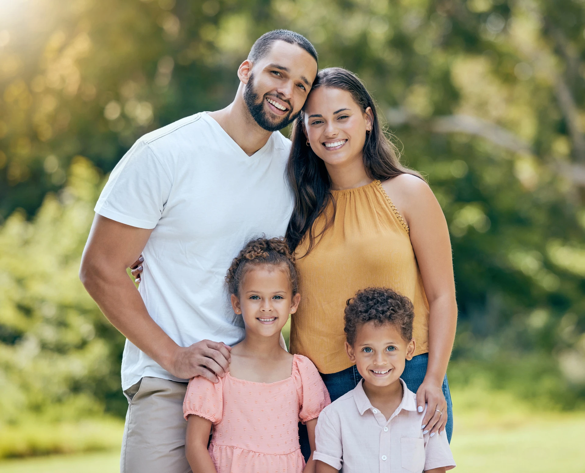 A man and woman stand together outdoors with two young children, all smiling at the camera in front of greenery.