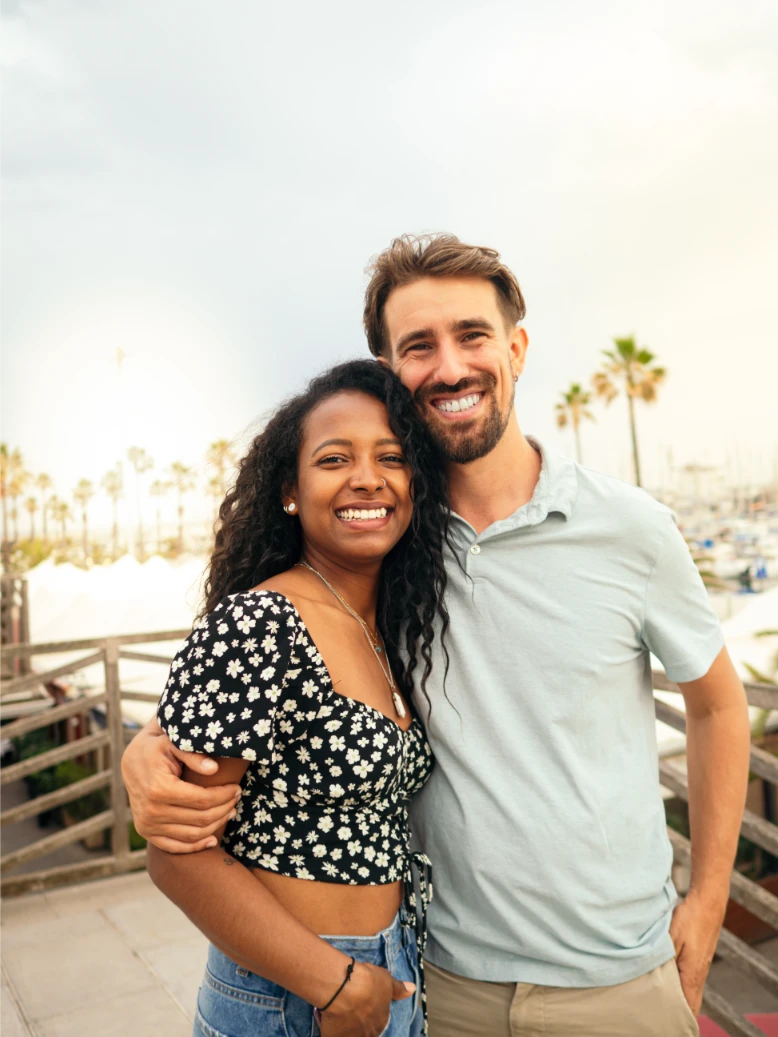 A smiling couple stands close together outdoors, with palm trees and a marina visible in the background.