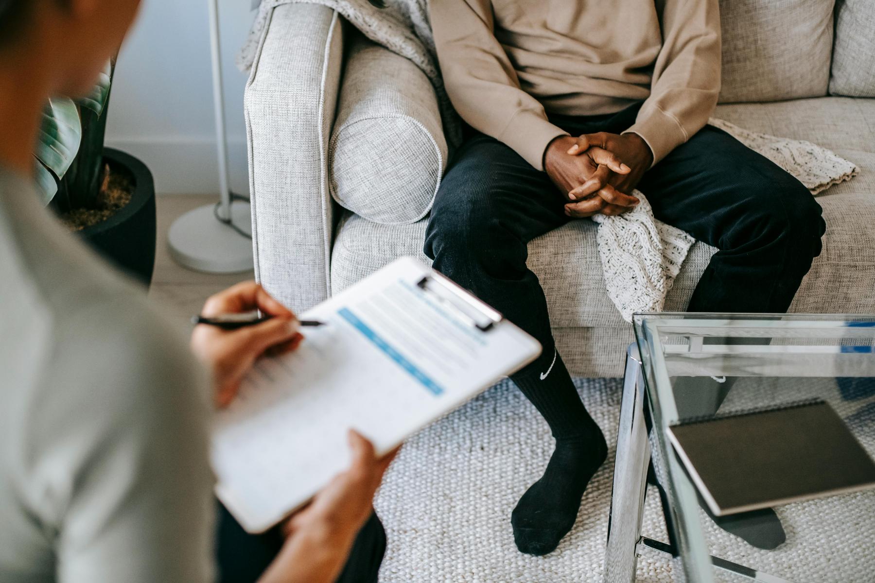 A person sits on a couch with hands clasped while another person takes notes on a clipboard during a conversation.