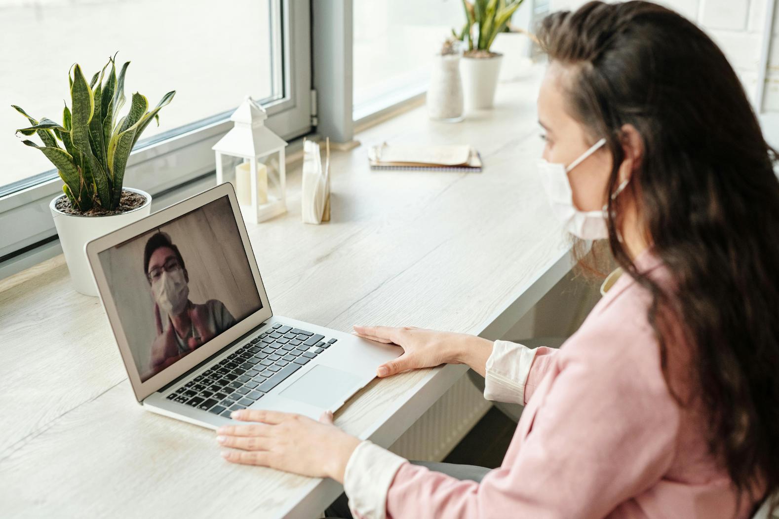 A woman wearing a mask sits at a desk and participates in a video call with another masked person on a laptop.
