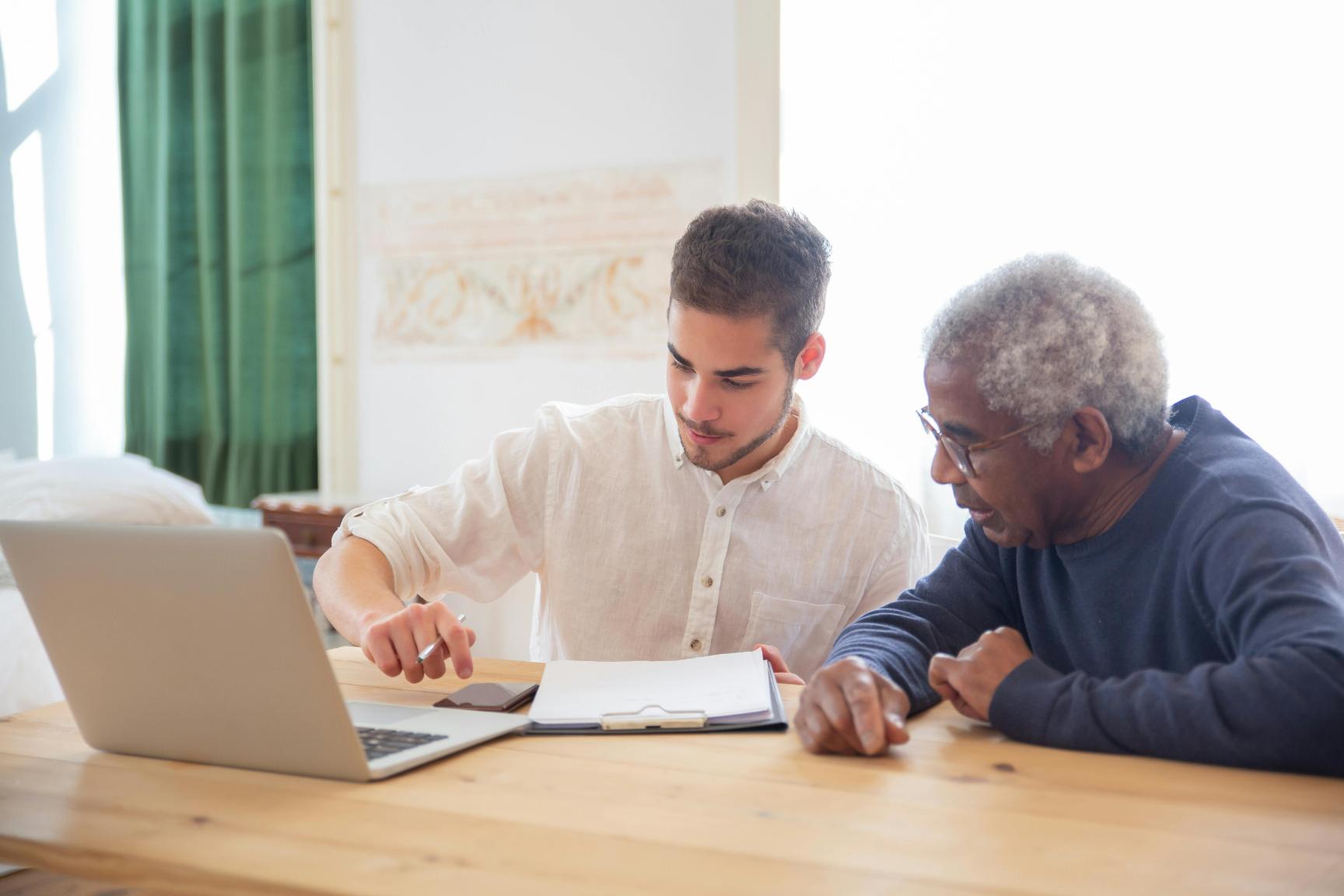 A young man shows an older man information on a laptop while discussing notes on a clipboard at a wooden table.
