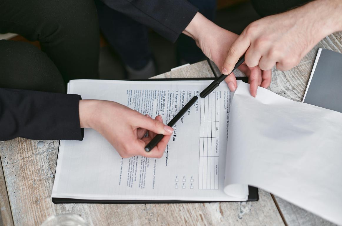 Two people review and point at a document on a clipboard, with one hand holding a pen and another hand turning a page on a wooden table.