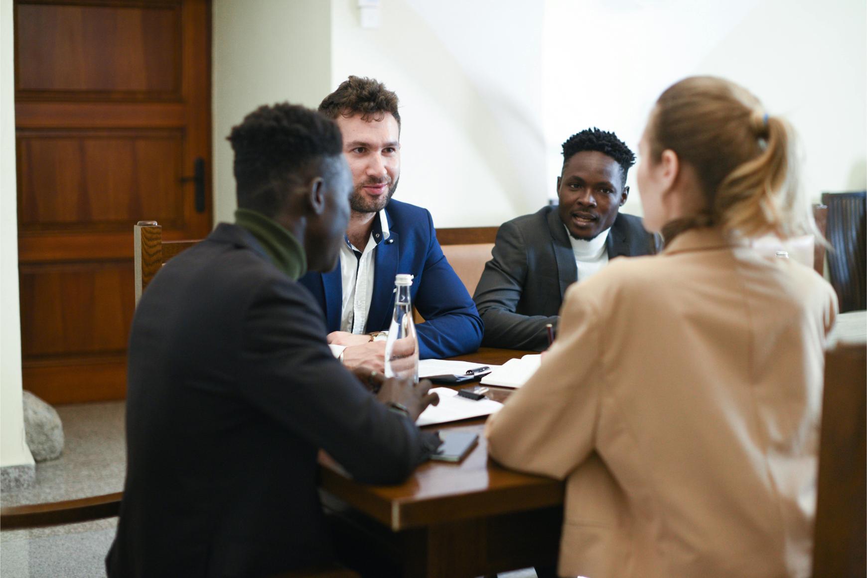 Four people sit around a table in a meeting, talking and taking notes, with water bottles and documents visible on the table.