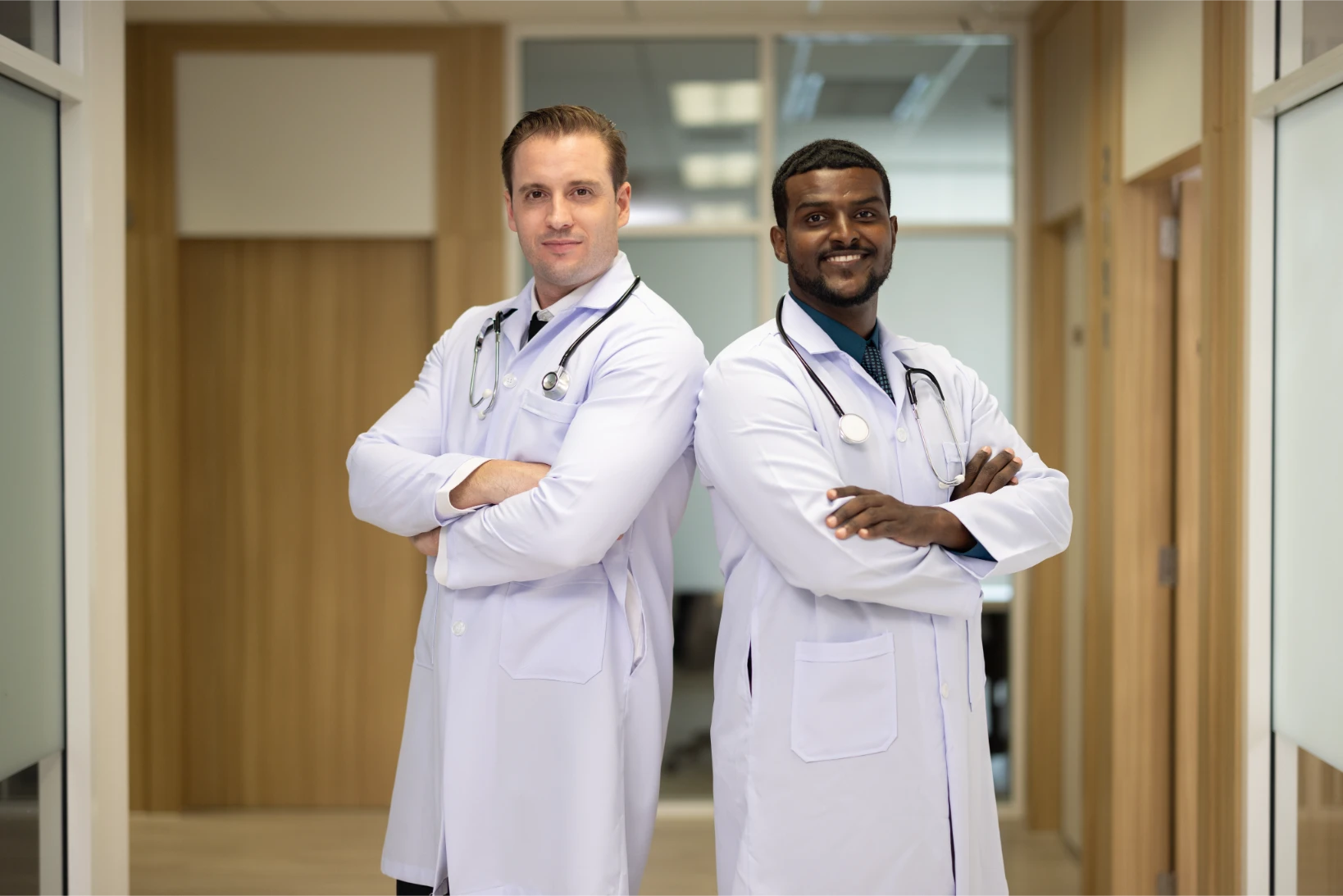 Two male doctors in white coats stand back-to-back with arms crossed in a hallway, both wearing stethoscopes and looking at the camera.